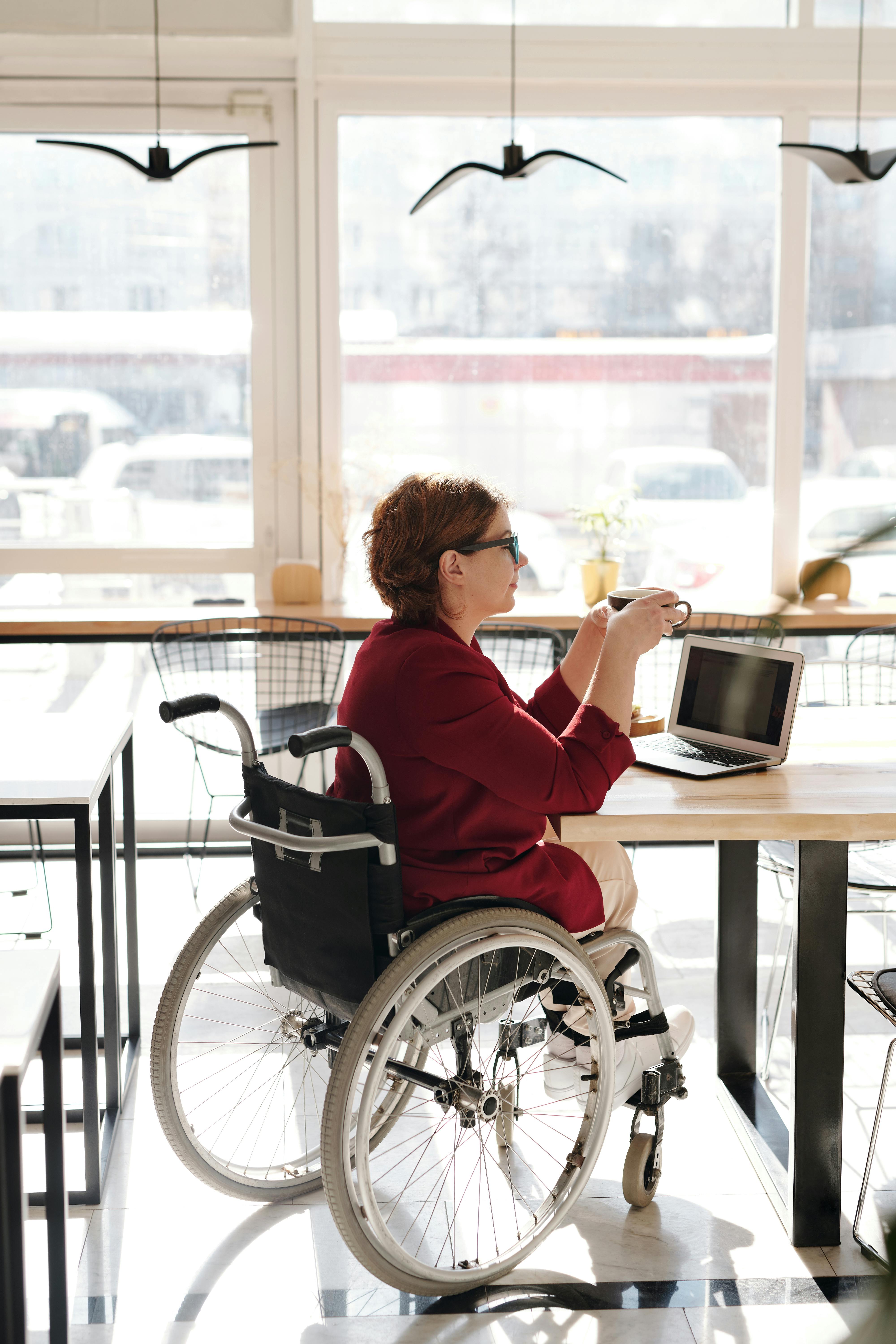 Person sitting in wheelchair with coffee and computer in cafe.